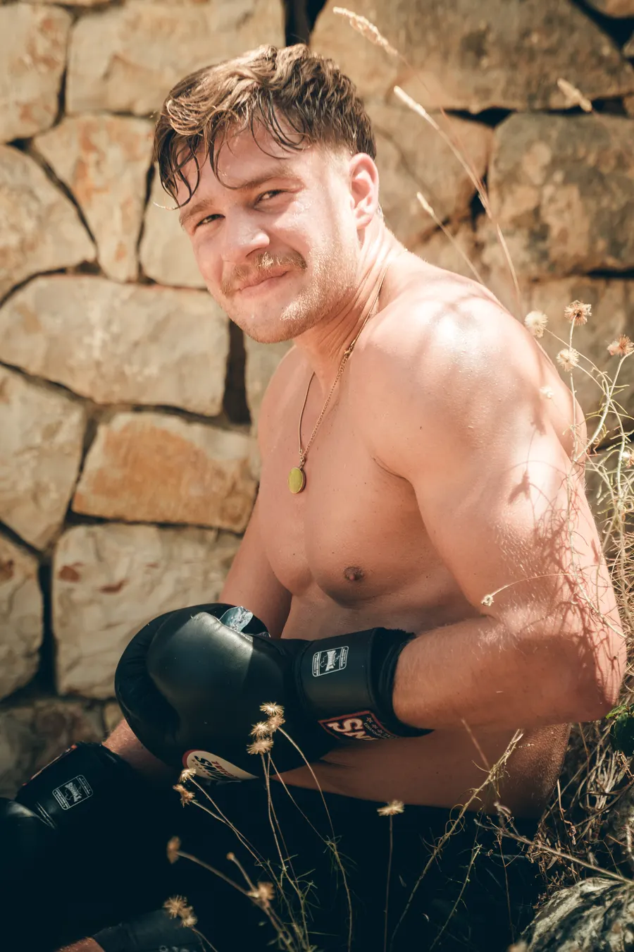 Ein Mann sitzt nach dem Sparring mit Boxhandschuhen auf einer Steinmauer und lacht.